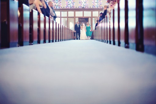 Bride walks down the aisle with her father and bridesmaid in a church