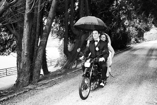 Newlyweds ride away on a motorcycle in wedding attire under an umbrella