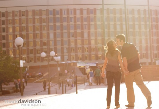 Couple holding hands kissing in front of a building in sunlight
