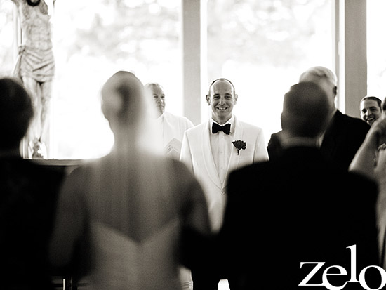 Groom in white suit smiles as bride walks down aisle