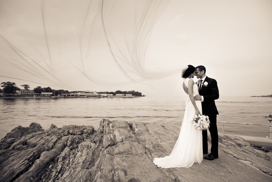 Bride and groom embrace on a rocky shore, her veil billowing