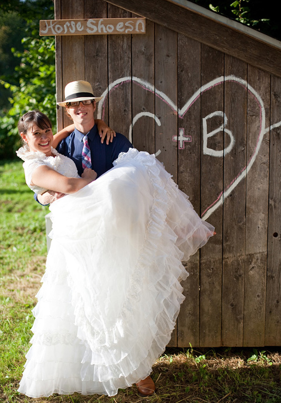 Groom carries smiling bride at their wedding with rustic backdrop