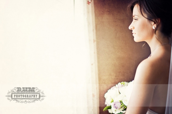 Bride in wedding dress looks out window, holding flowers