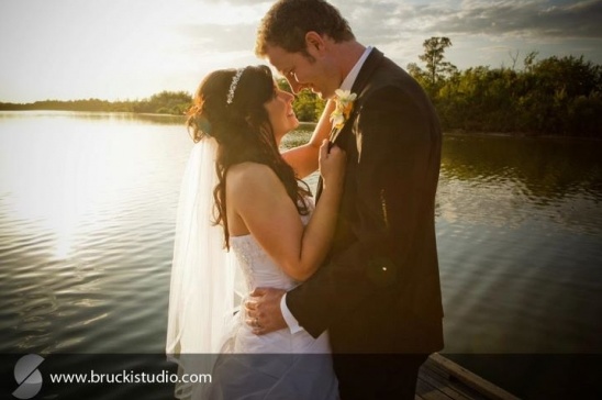 Bride and groom embrace by lake at sunset wedding
