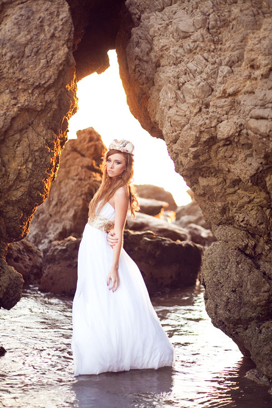 Woman in white dress and crown stands in ocean cove