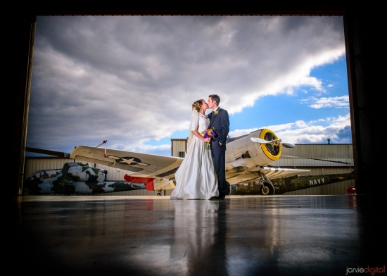 Wedding couple kisses in front of old navy plane under cloudy sky