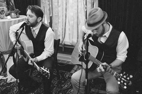 Two musicians in formal attire play guitars and sing together