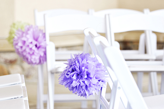 Lavender paper pom-poms adorn white wedding chairs