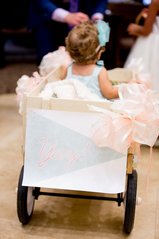 A cute flower girl in a wagon at a wedding with a Yay! sign