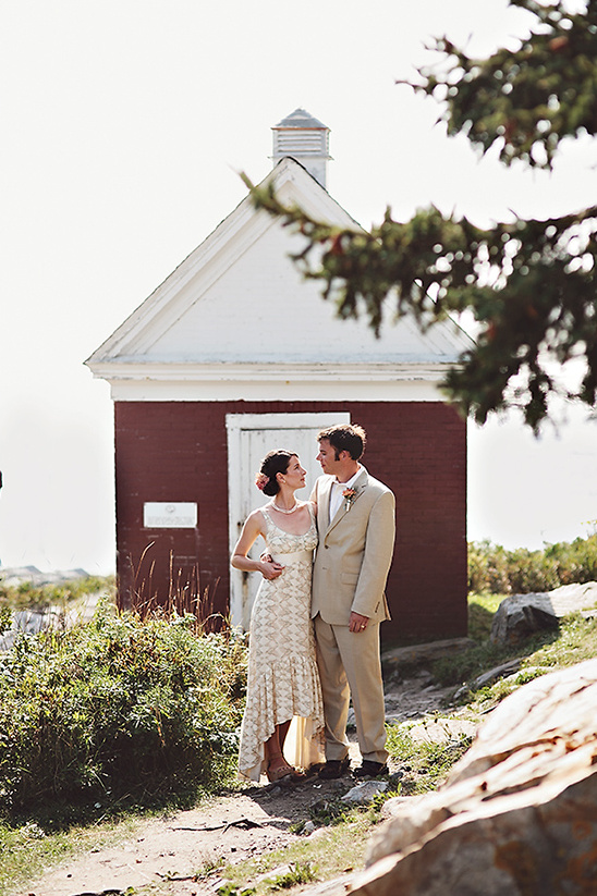 Bride and groom pose by a small red building