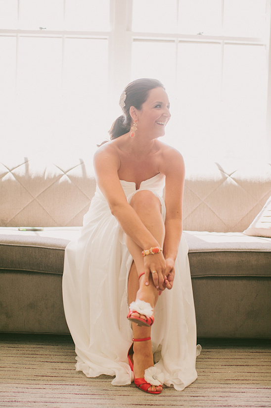Bride smiling in a white dress with red floral sandals, waiting