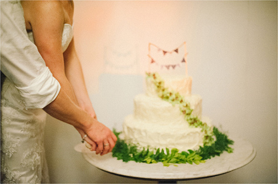 Bride and groom cut wedding cake adorned with green foliage