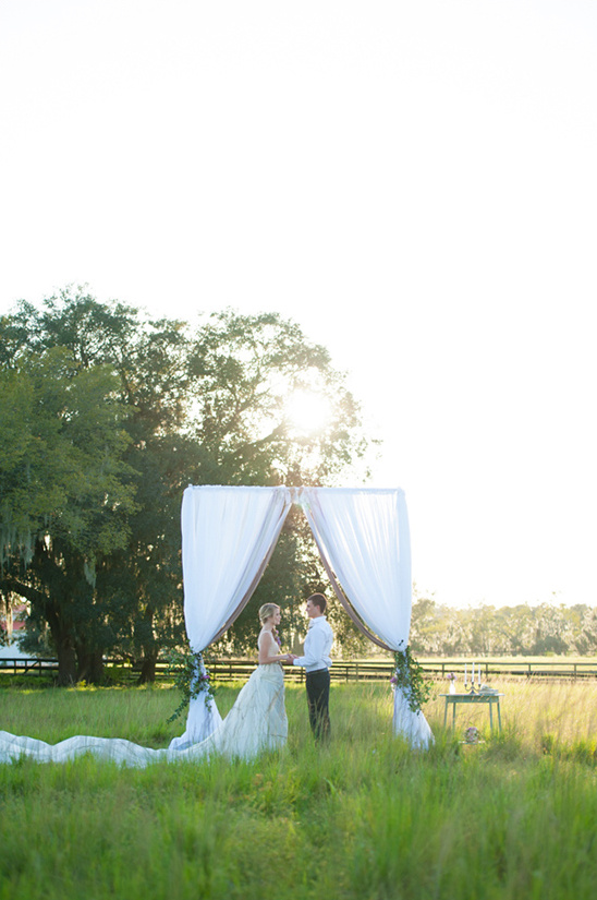 Couple exchanging vows under a wedding arch in a sunny field