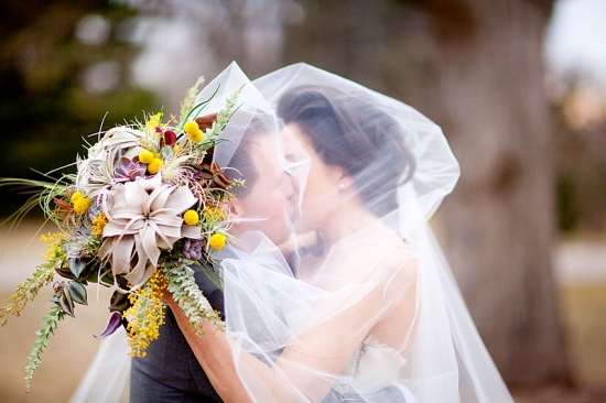 Bride and groom kiss under veil, holding bouquet