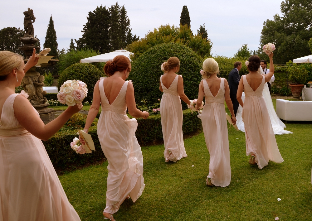 Bridesmaids in pink gowns walk across a garden lawn