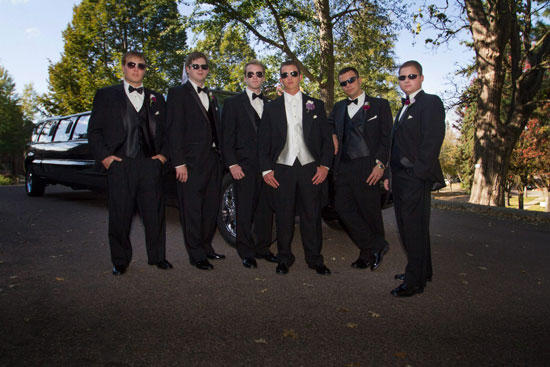 Groomsmen in tuxedos pose by limousine