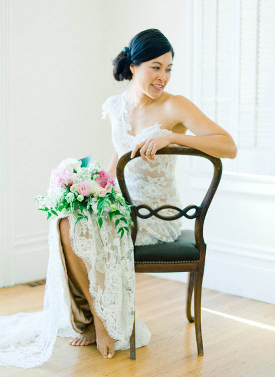 Bride in lace gown sits with bouquet on chair smiling