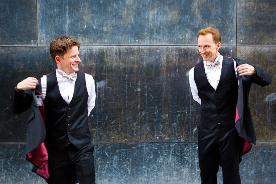 Two men in black tuxedos against wall