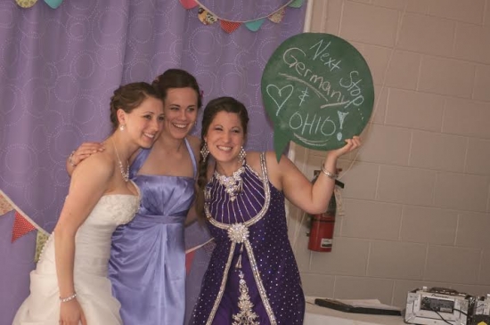 Three friends at a wedding, holding a sign: Next stop Germany & Ohio!