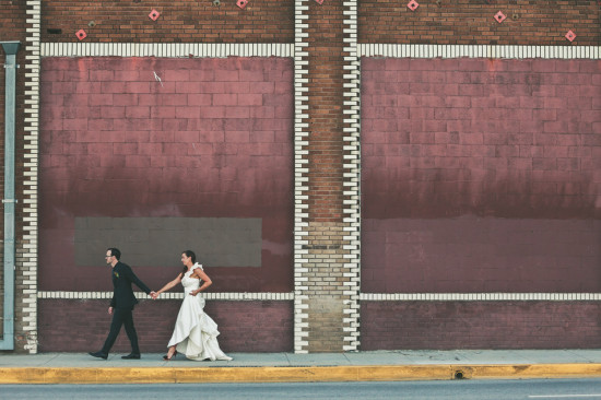 Bride and groom stroll past urban brick wall backdrop