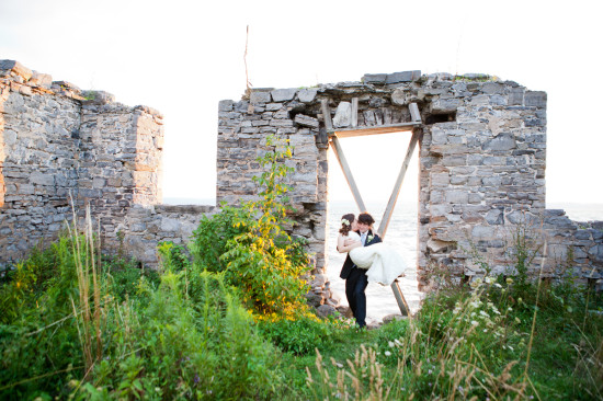 Groom carries bride kissing in stone ruins by water