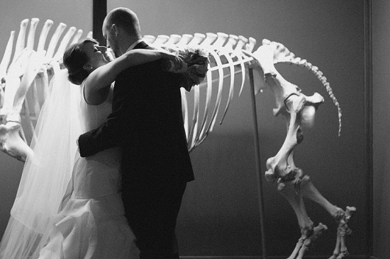 Newlyweds embrace with dinosaur skeleton backdrop at wedding