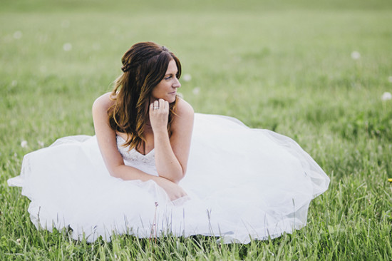 Bride in white gown sits contemplatively in grassy field