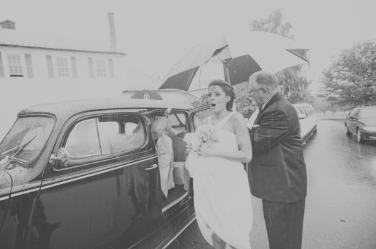 Bride exiting car in the rain, being helped with an umbrella