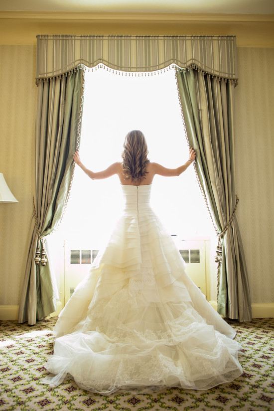 Bride in wedding dress looking out the window with open curtains