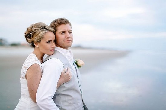 Wedding couple embrace on the beach, looking towards the horizon