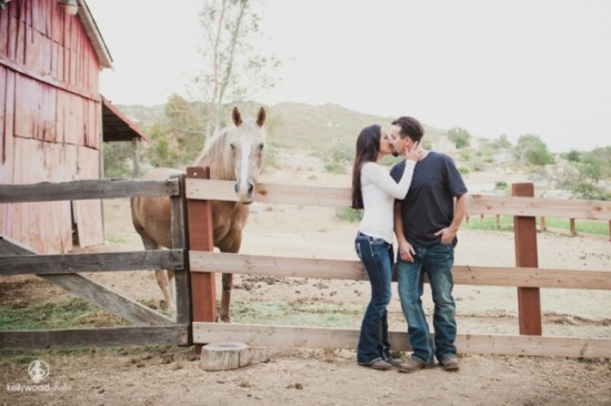 Couple kissing by a fence with a horse and a red barn in background