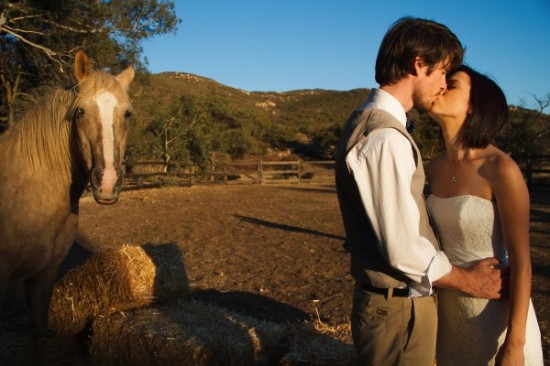 Newlyweds sharing a kiss as a horse curiously watches from the side