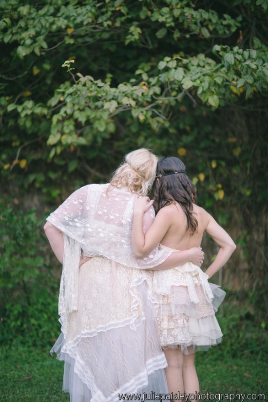 Two women in delicate lace dresses embrace, back to the camera, under green trees