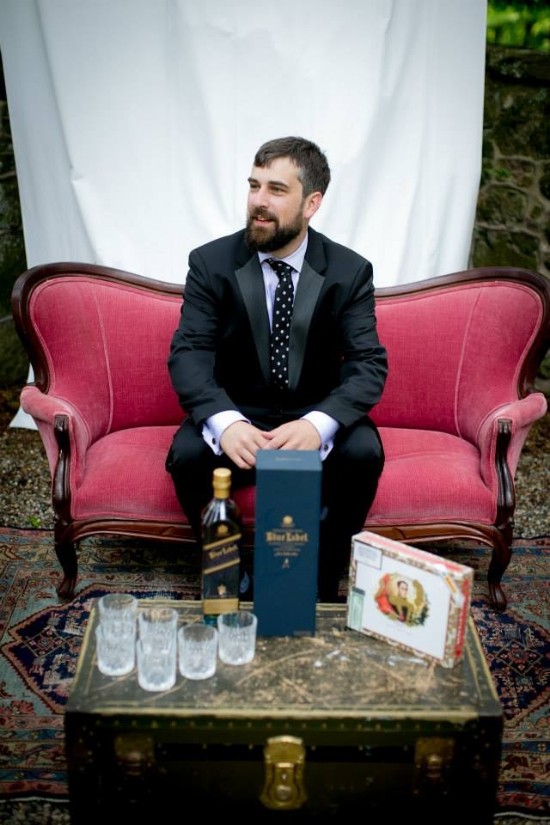 Groom in tuxedo sits on a red sofa with whiskey and cigars