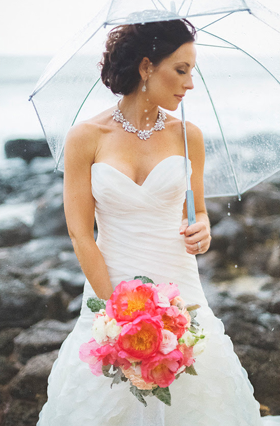 Bride in white dress holding coral bouquet under umbrella