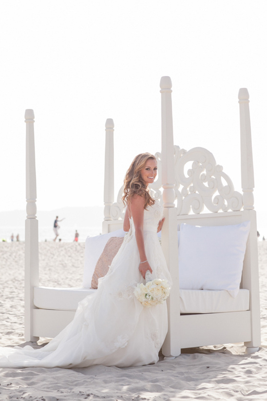 Bride in a white dress poses on a beach beside ornate bed