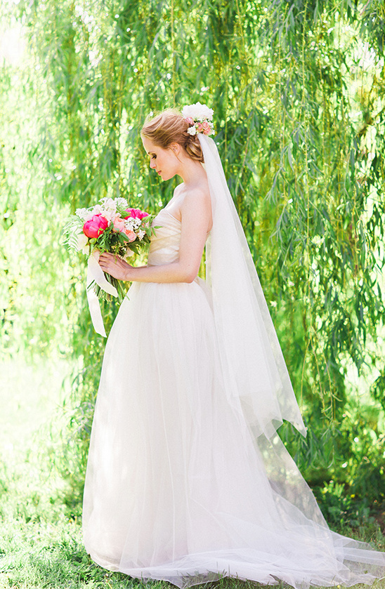 Bride in white gown holding bouquet by willow tree