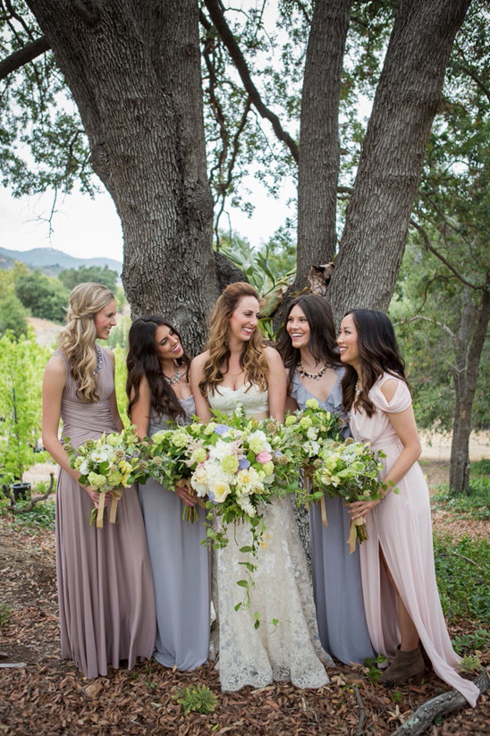 Bride and bridesmaids in pastel gowns smiling near a tree