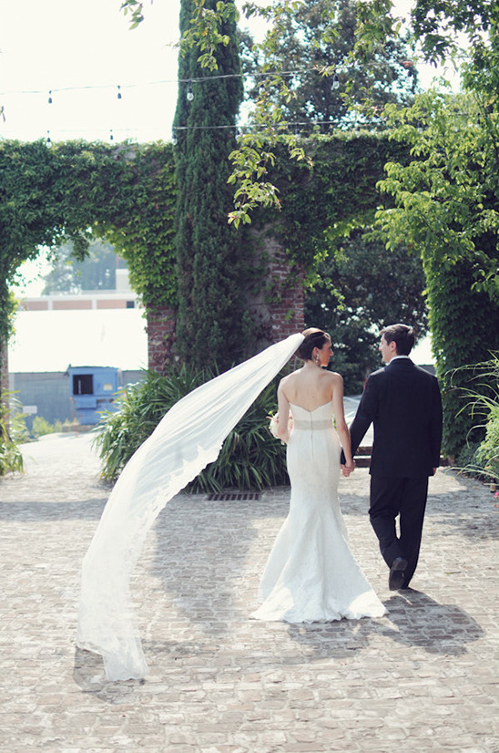 Bride and groom walk holding hands with veil flowing