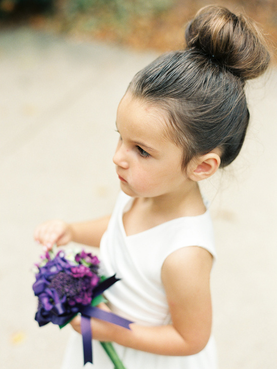 Elegant flower girl in white dress holds purple bouquet