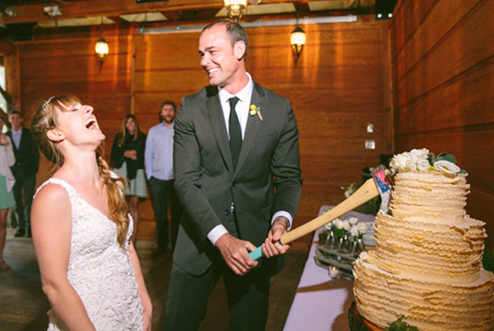 Bride and groom laughing while cutting the wedding cake with an axe