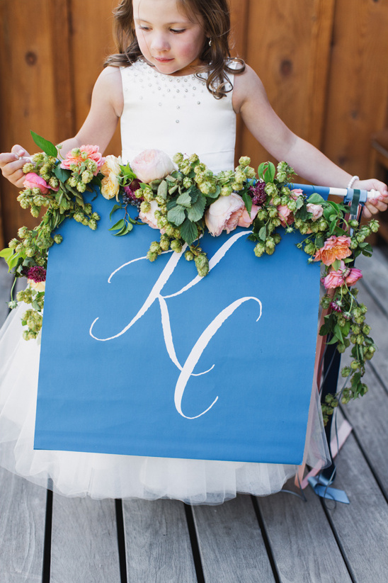 Flower girl holding a blue sign with initials and floral garland