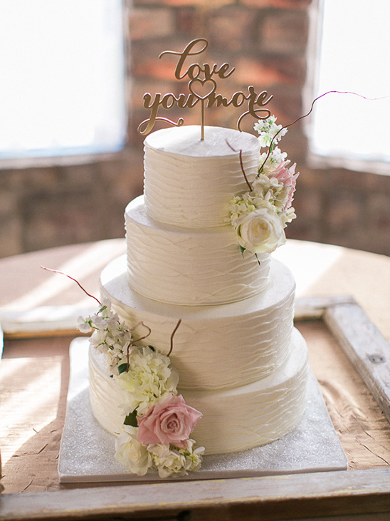 Elegant four-tiered wedding cake decorated with flowers and a Love You More topper
