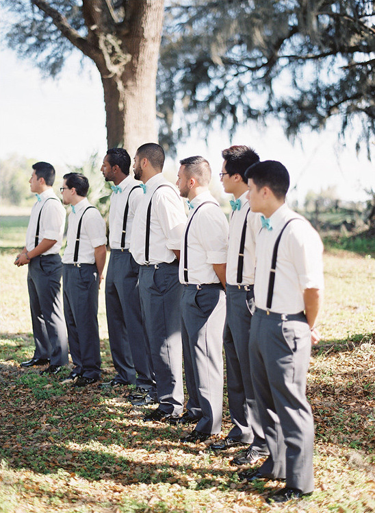 Groomsmen line up for a wedding ceremony outdoors with suspenders