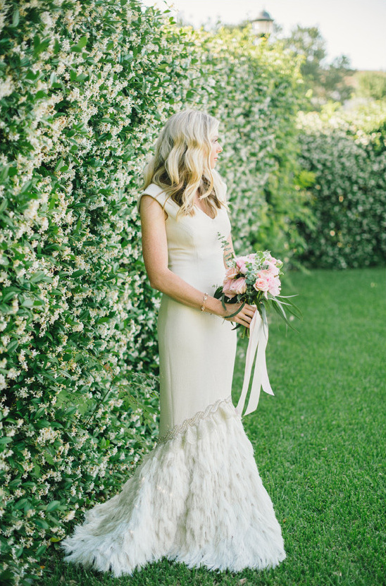 Bride in a feathered gown holds bouquet by flower hedge