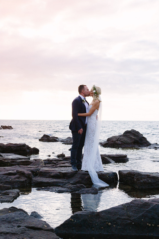 Bride and groom share a kiss on rocky beach at sunset