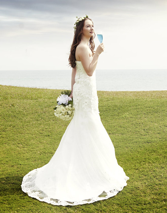 Bride in gown near ocean holding bouquet and drink