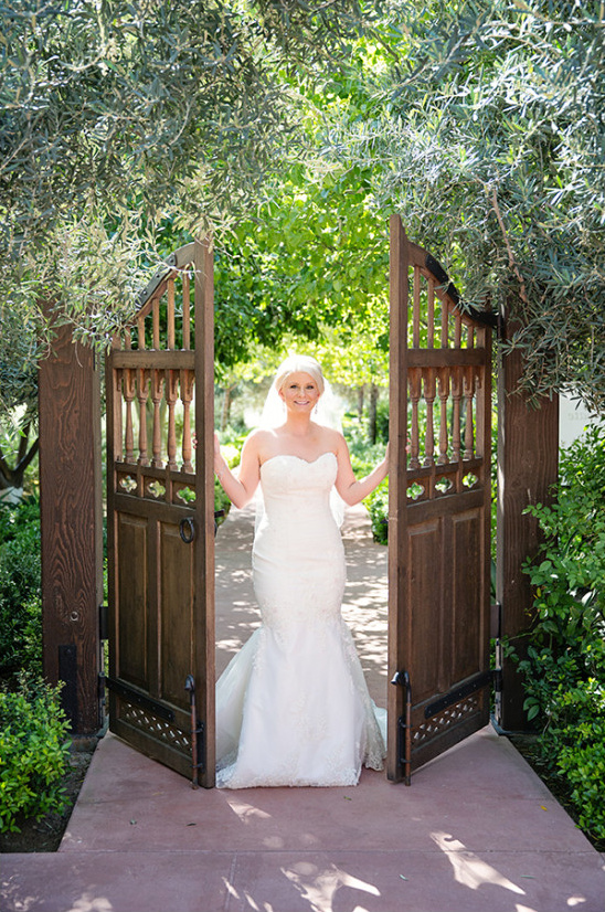 Bride in wedding dress stands at wooden garden gate