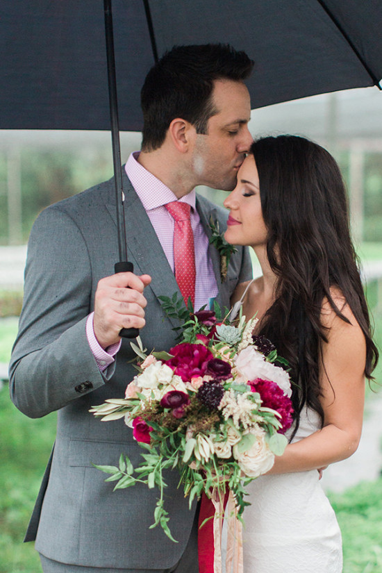 Groom kisses bride under umbrella, holding bouquet