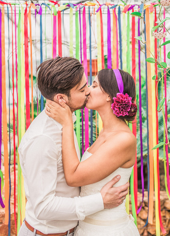 Newlyweds share a kiss in front of a colorful ribbon backdrop